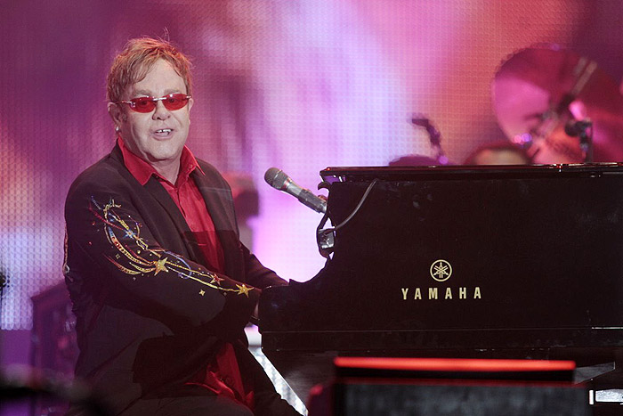 Elton John sentado ao piano e com os tradicionais óculos coloridos