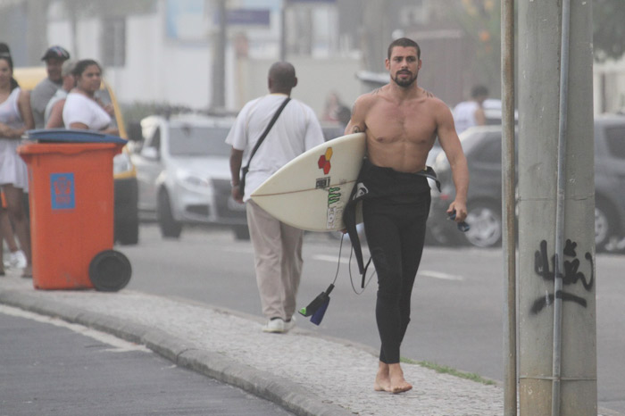 Cauã Reymond anda por orla com sorriso no rosto e prancha nas mãos