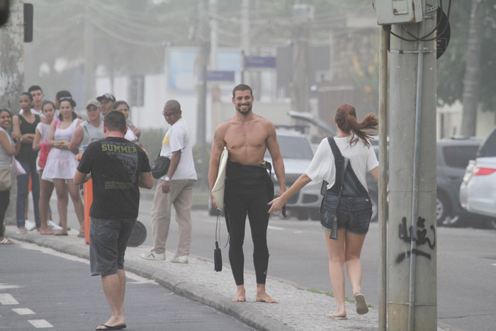 Cauã Reymond anda por orla com sorriso no rosto e prancha nas mãos