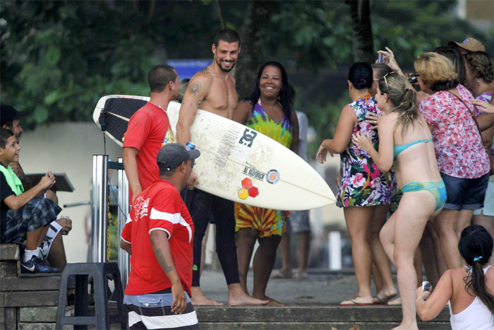 Cauã Reymond anda por orla com sorriso no rosto e prancha nas mãos