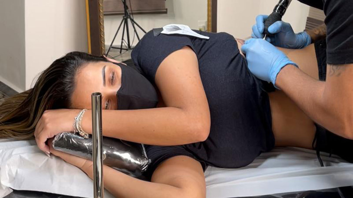 Ivy Moraes deitada na maca enquanto o tatuador faz a tatuagem