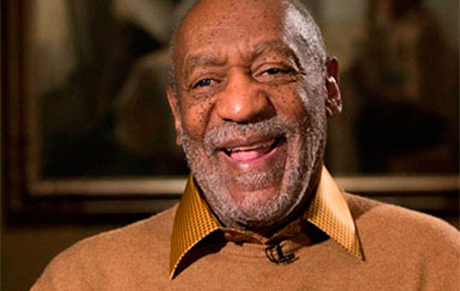 Bill Cosby de blusa cor de caramelo, sentado, sorrindo