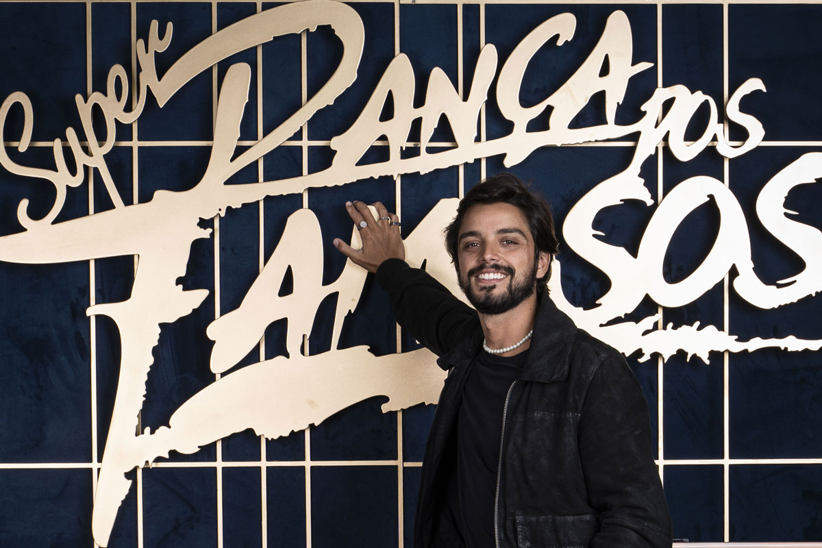Rodrigo Simas sorrindo com a logo do Super Dança dos Famosos atrás