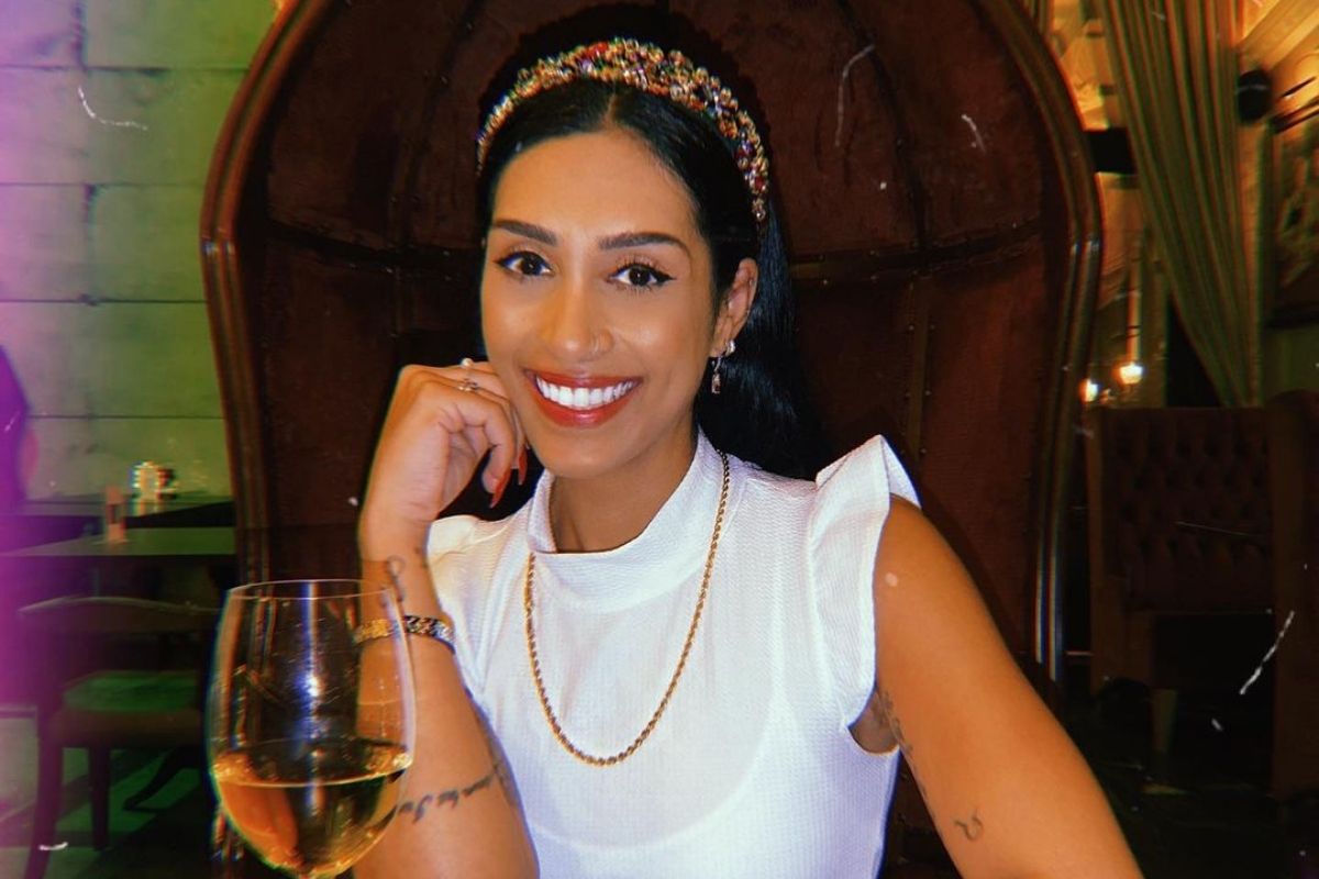 Foto de Amanda Djehdian sorrindo, usando uma tiara na cabeça e com uma taça de vinho a sua frente