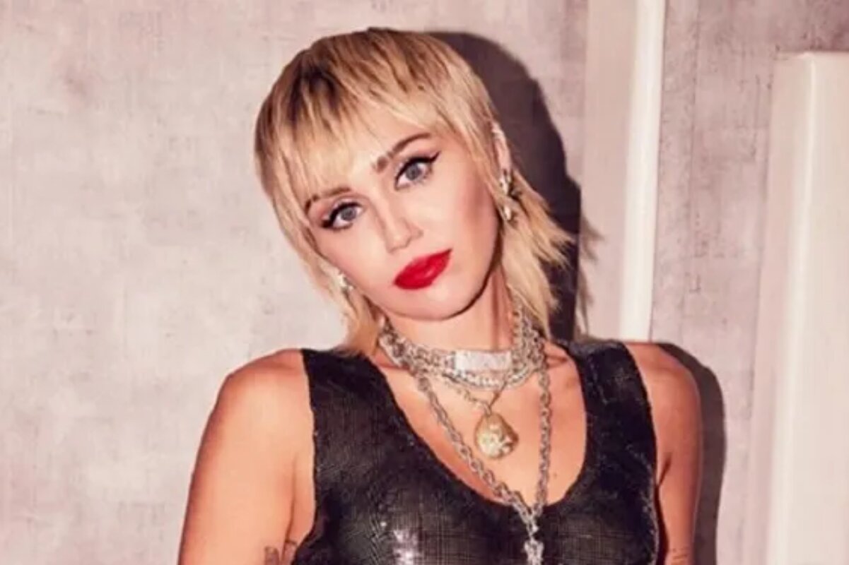 Miley Cyrus de cabelo curto e blusa transparente preta, batom vermelho