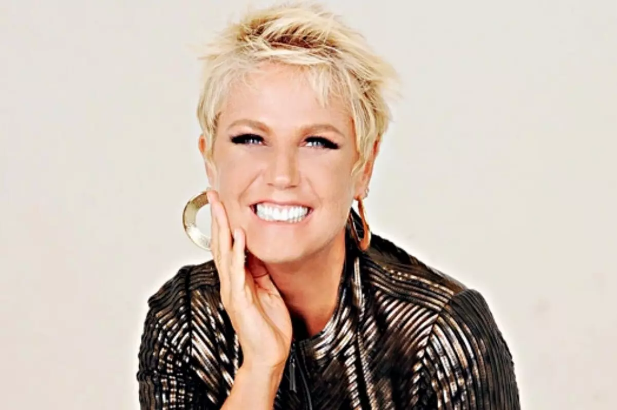 Xuxa Meneghel sorrindo, de roupa preta brilhante, com a mão direita no queixo