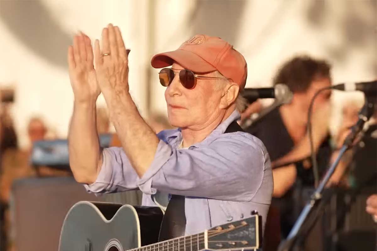 Paul Simon de óculos escuro, aplaudindo, durante aparição surpresa em festival nos EUA