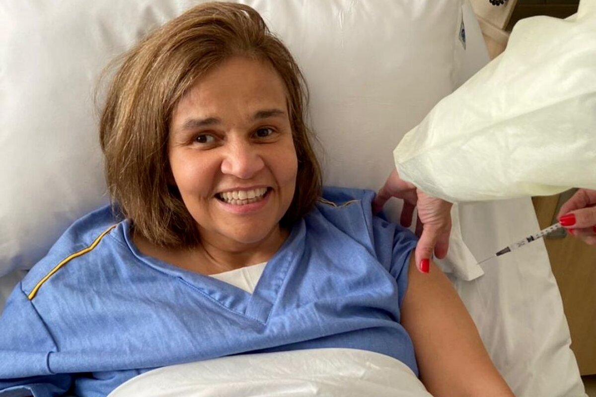 Claudia Rodrigues na cama do hospital, de roupão azul, sorrindo, tomando injeção