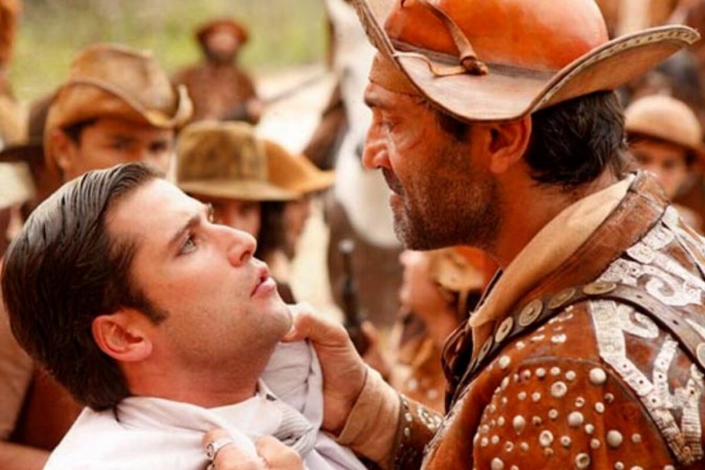 Bruno Gagliasso de terno branco,sendo puxado pelo colarinho por  Domingos Montagner, que está de roupa de cangaceiro, em cena da novela Cordel Encantado