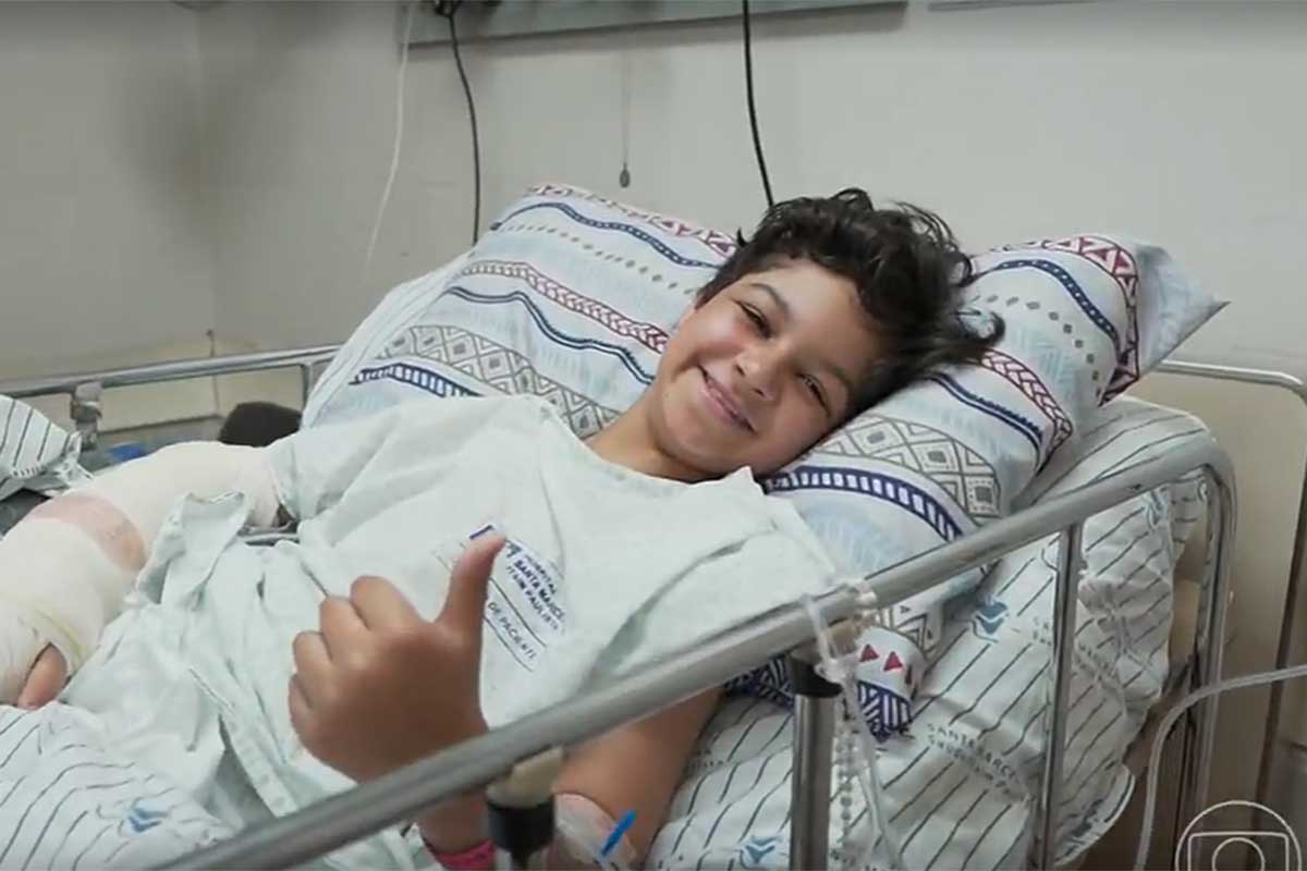 Gustavo Corasini, na cama do hospital, fazendo sinal de positivo e sorrindo
