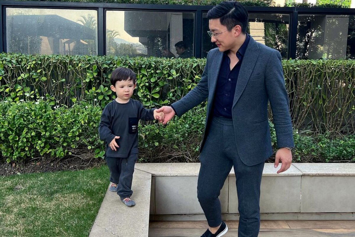 Pyong Lee de calça e blazer escuro, segurando a mão do filho, Jake, que anda sob uma área livre