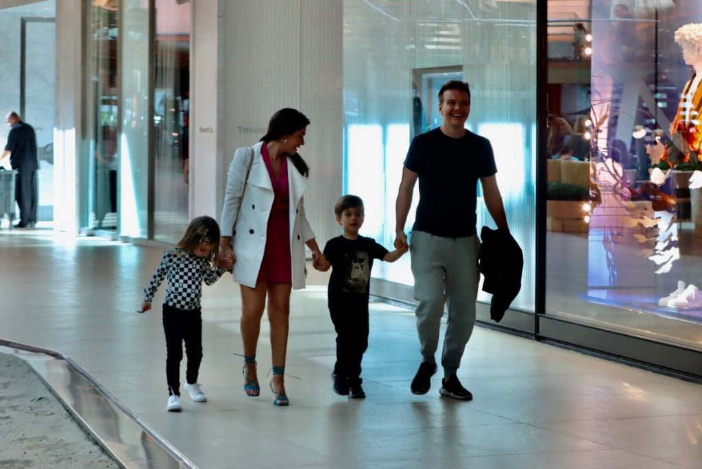 Michel Teló de camisa preta, segurando a jaqueta, Thais Fersoza de casaco branco e vestido rosa, Melinda e Theodoro passeando no corredor do shopping