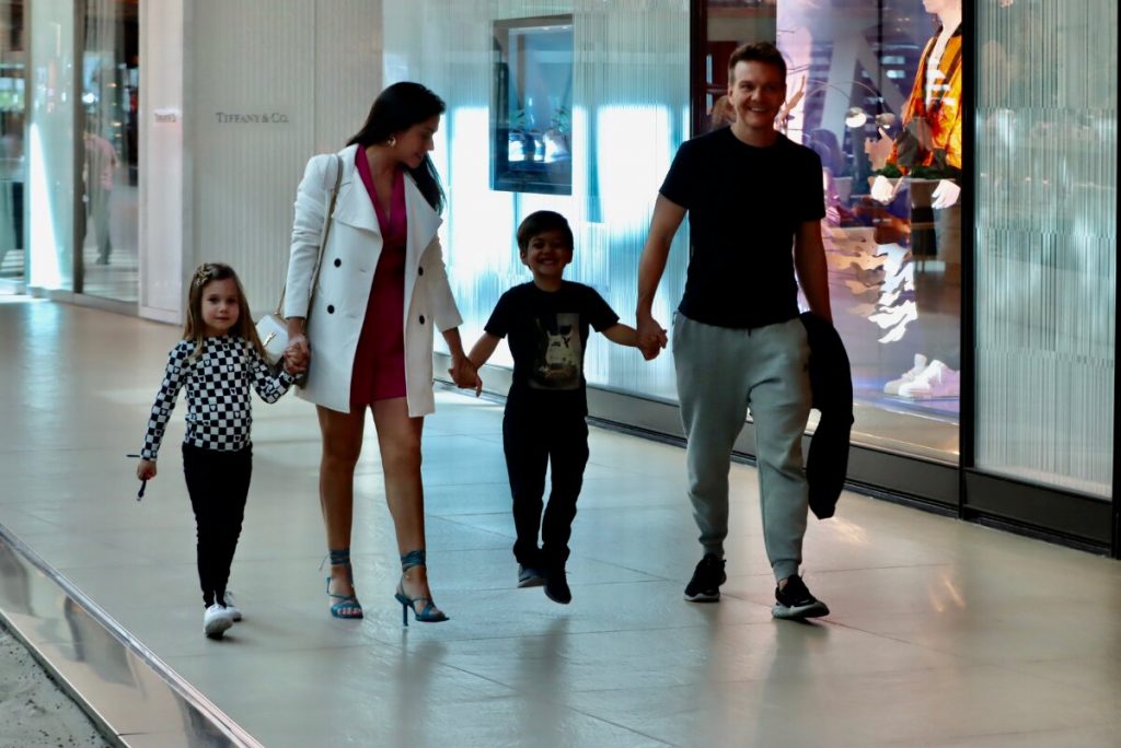 Michel Teló de camisa preta, segurando a jaqueta, Thais Fersoza de casaco branco e vestido rosa, Melinda e Theodoro passeando no corredor do shopping