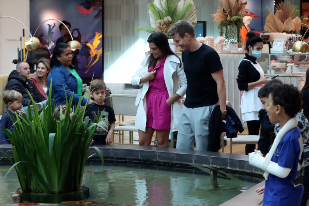 Michel Teló de camisa preta, segurando a jaqueta, Thais Fersoza de casaco branco e vestido rosa, Melinda e Theodoro diante de uma fonte, no shopping