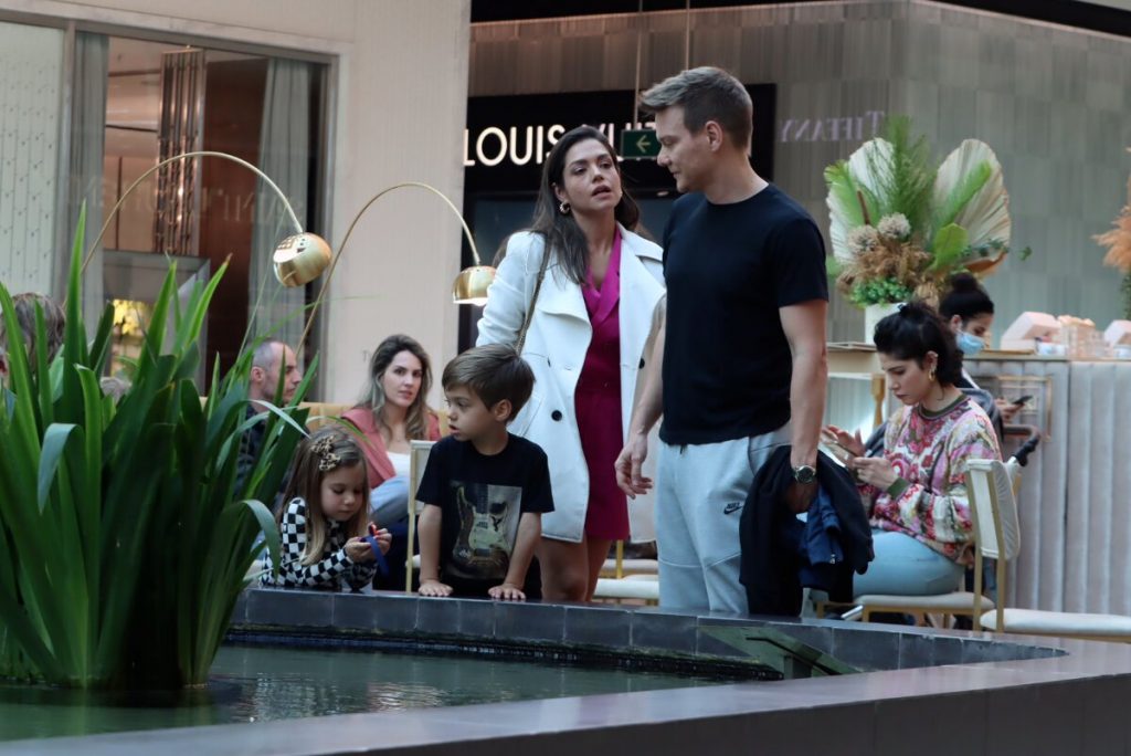 Michel Teló de camisa preta, segurando a jaqueta, Thais Fersoza de casaco branco e vestido rosa, Melinda e Theodoro diante de uma fonte, no shopping