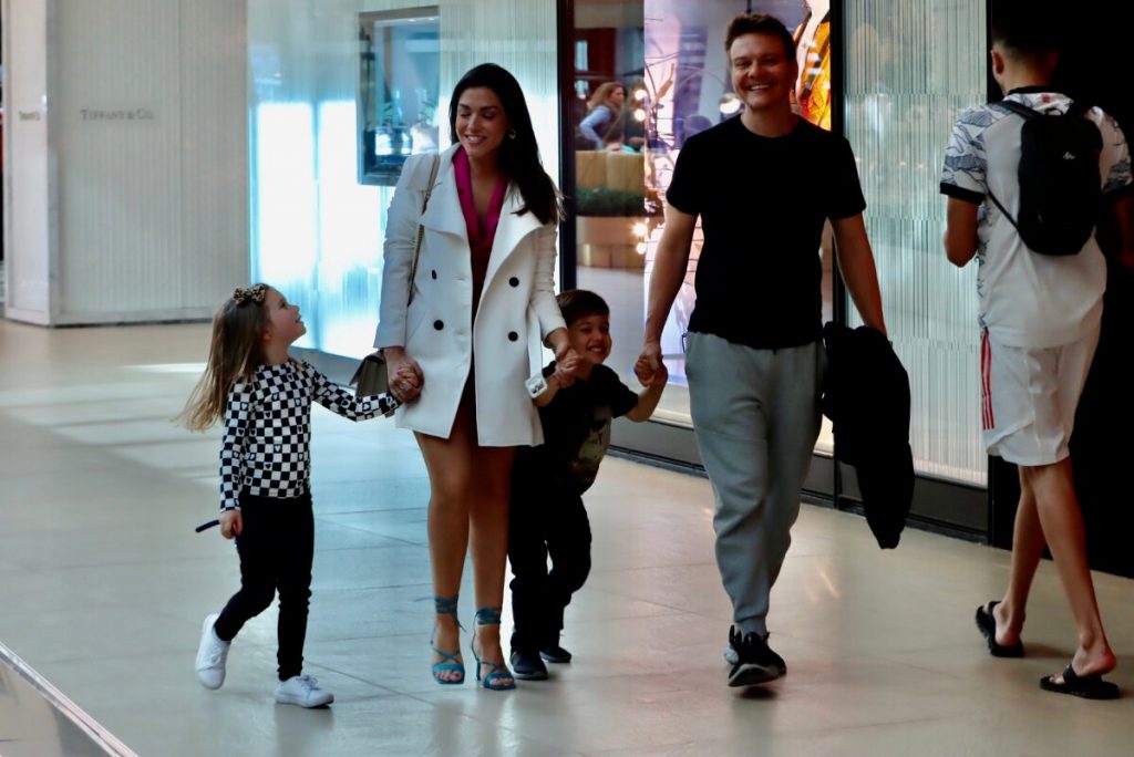 Michel Teló de camisa preta, segurando a jaqueta, Thais Fersoza de casaco branco e vestido rosa, Melinda e Theodoro passeando no corredor do shopping