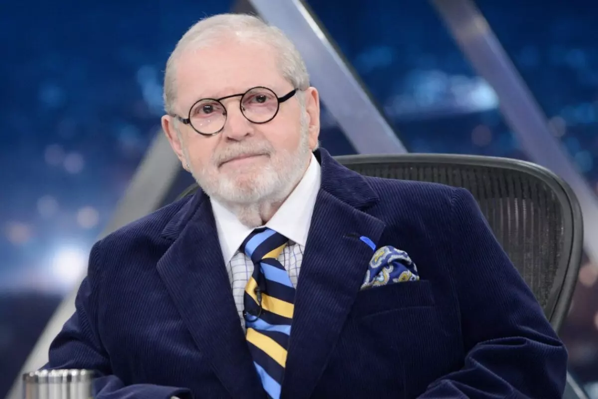 Jô Soares sentado no cenário do programa "Jô Onze e Meia", de terno preto, óculos, olhando para a câmera/