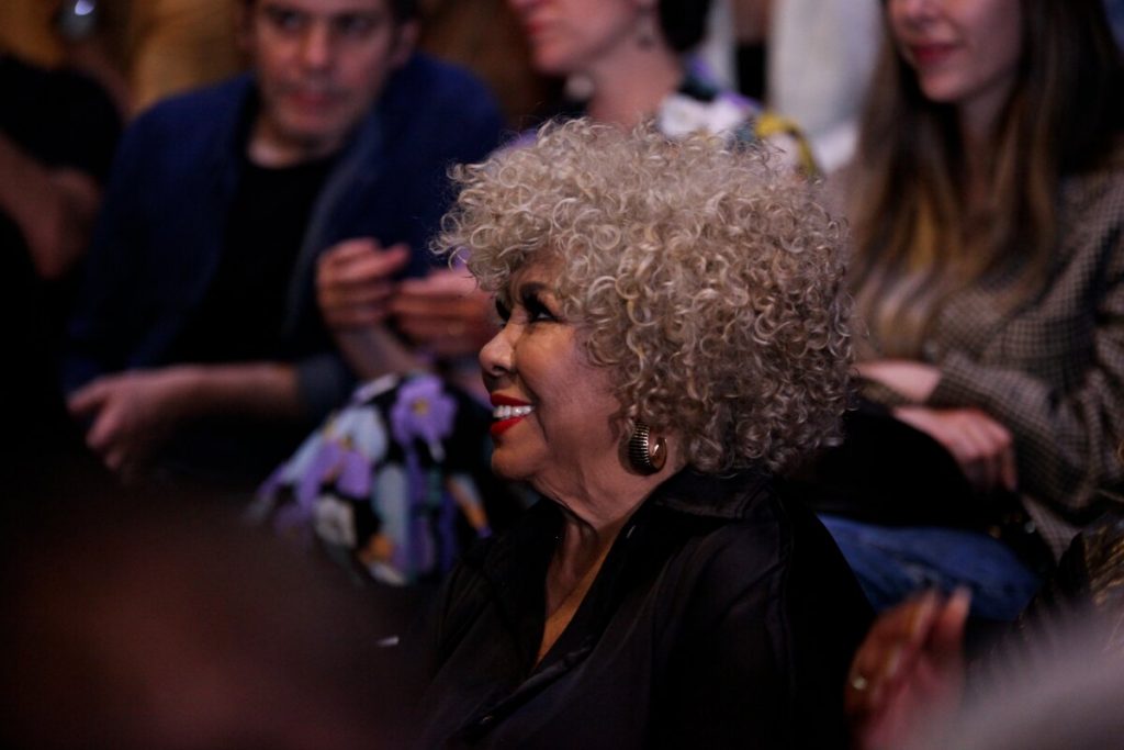 Alcione sorrindo de preto, sentada na plateia do teatro