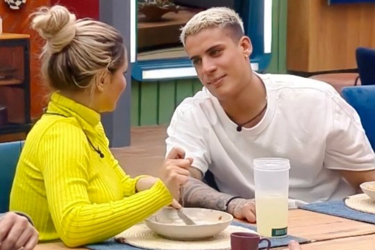 Deolane de blusa de gola alta o amarela, olhando pra Tiago Ramos, que veste camisa branca, ambos sentados à mesa