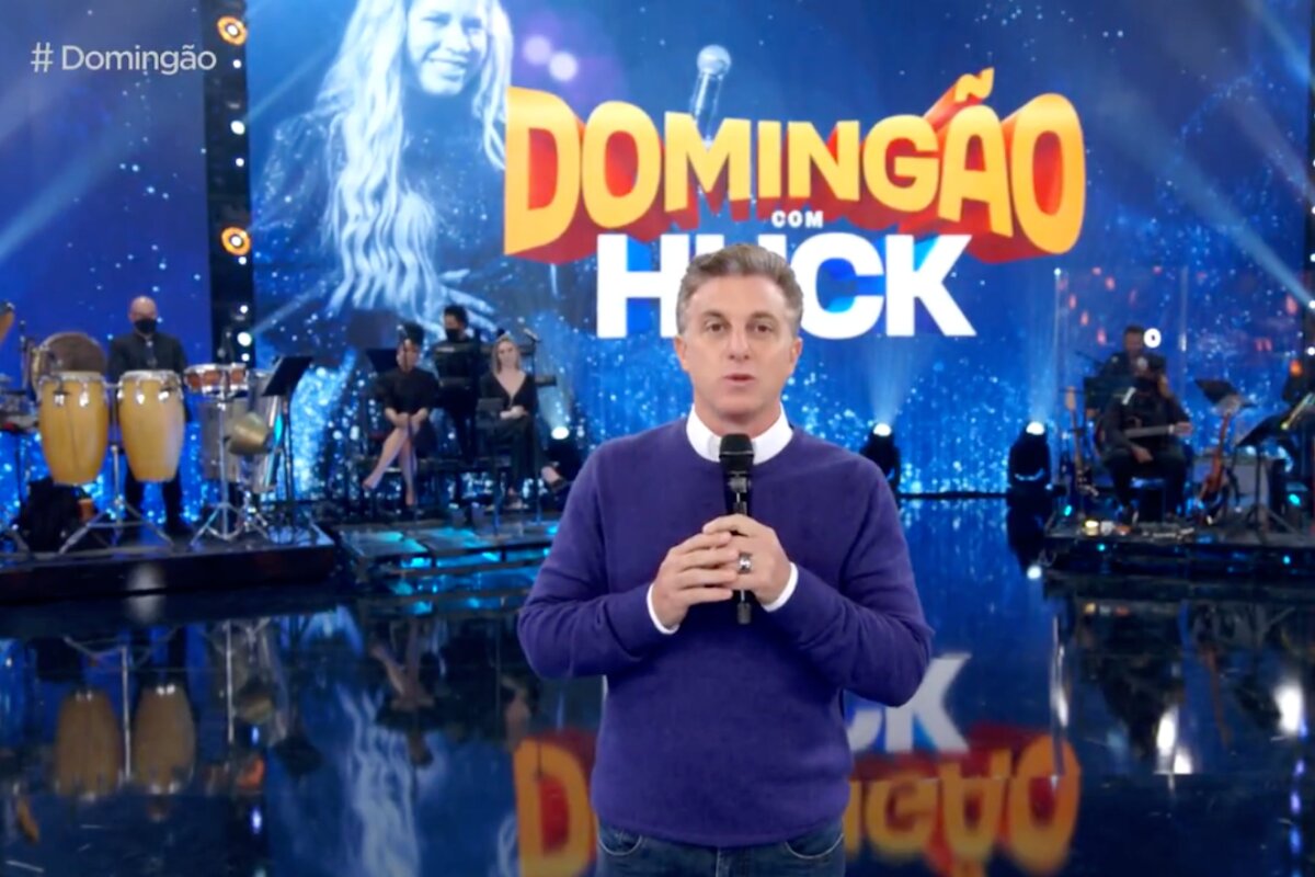 Luciano Huck de blusa de manga comprida azul, mãos cruzadas, no palco do "Domingão com Huck"