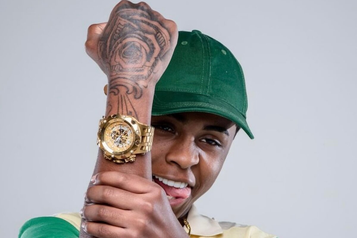 MC Meno K de boné verde, fazendo gesto segurando o braço pra cima, exibindo relógio dourado
