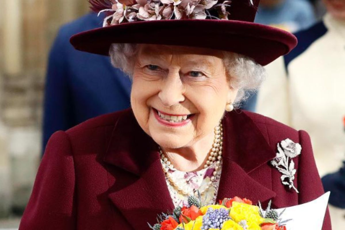 Rainha Elizabeth II com roupa e chapéu bordô, segurando flores amarelas, sorridente
