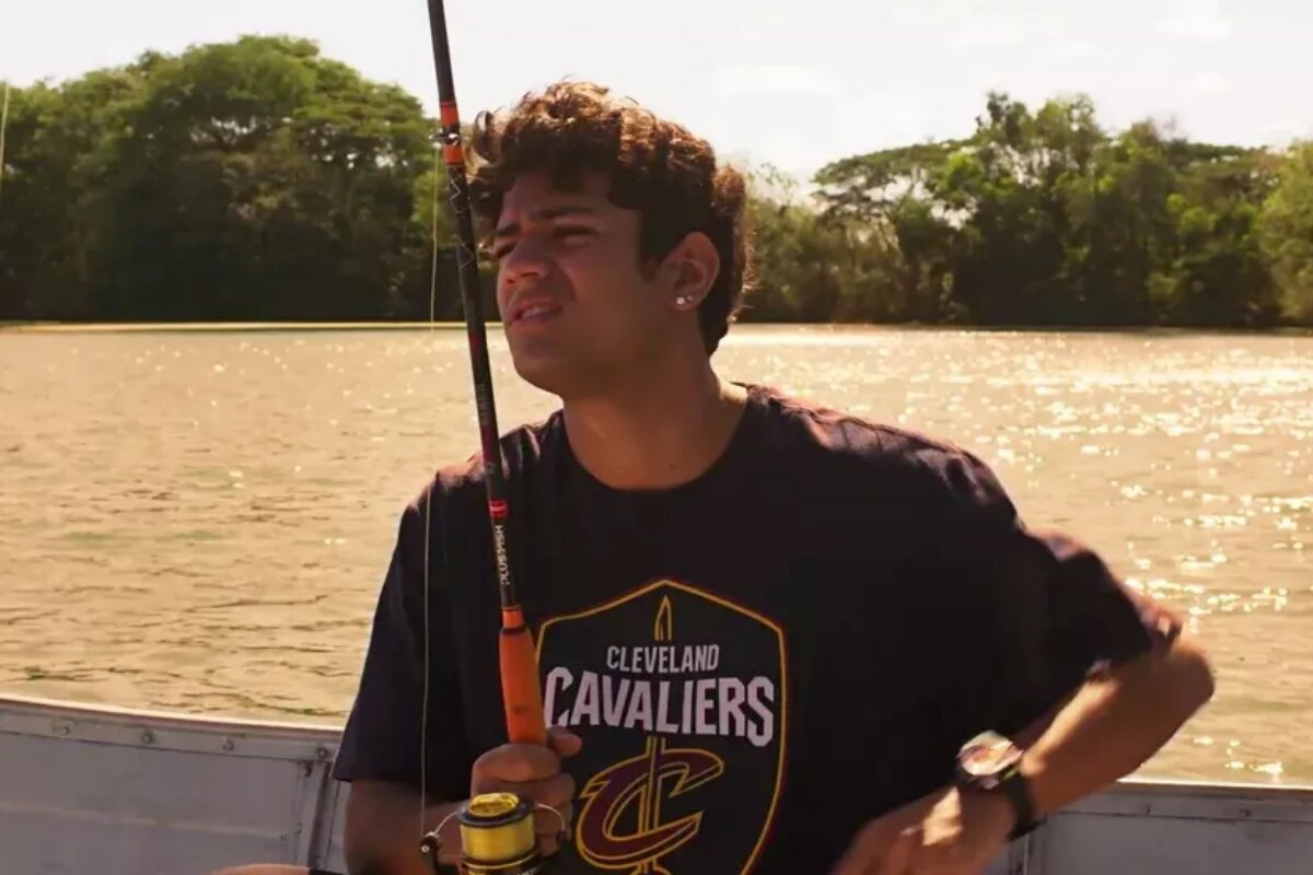 Renato (Gabriel Santana) de camisa preta, segurando uma vara de pesca, no barco