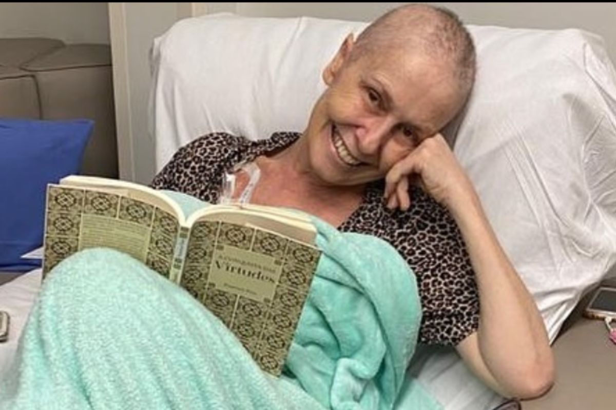 Susana Naspolini na cama do hospital, internada, segurando um livro e sorrindo