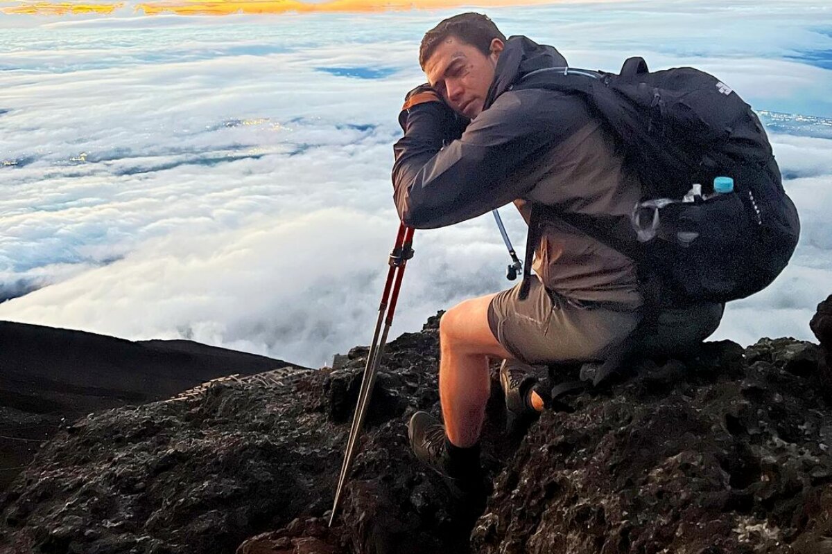 Whindersson Nunes no alto do Monte Fuji, ce casado, bermuda e mochila nas costas, sentado