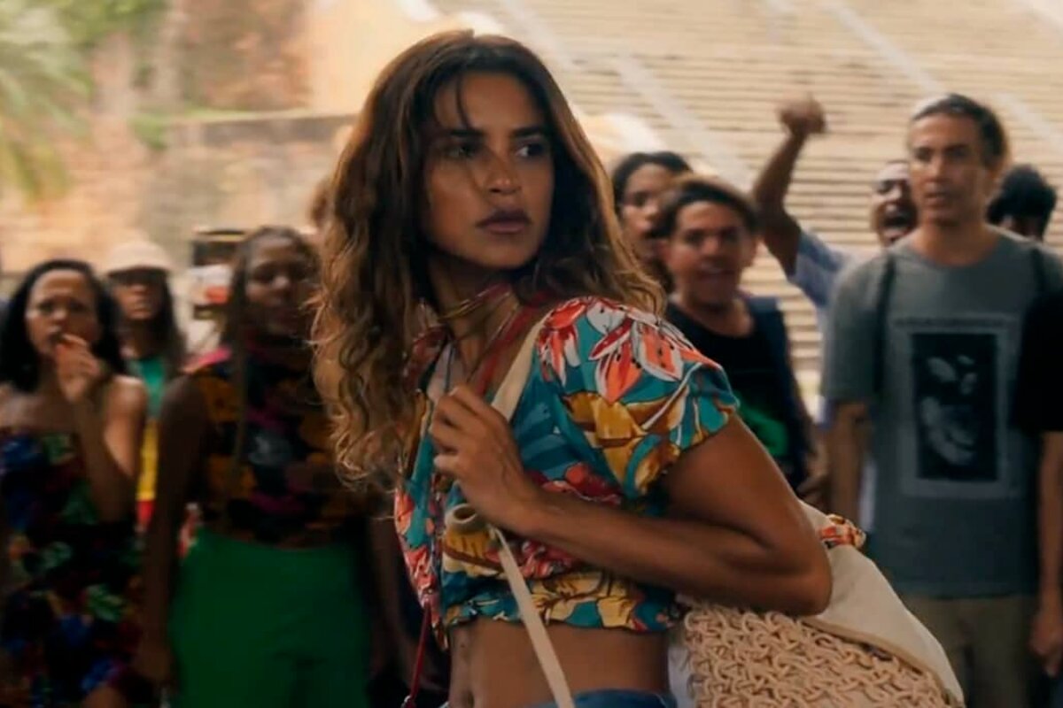 Brisa (Lucy Alves) de cropped estampado, barrida de fora, com uma bolsa à tiracolo, em meio a varias pessoas