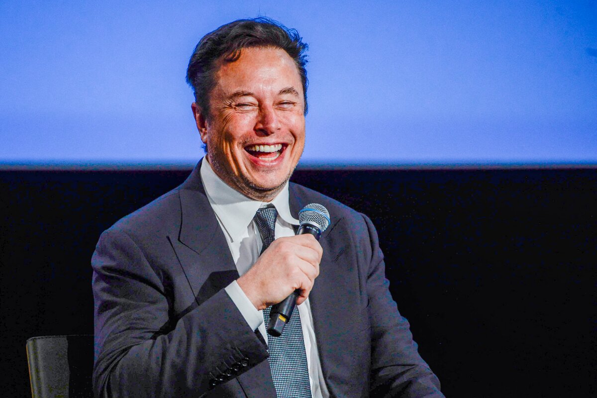 Elon Musk sorrindo, de terno, segurando microfone