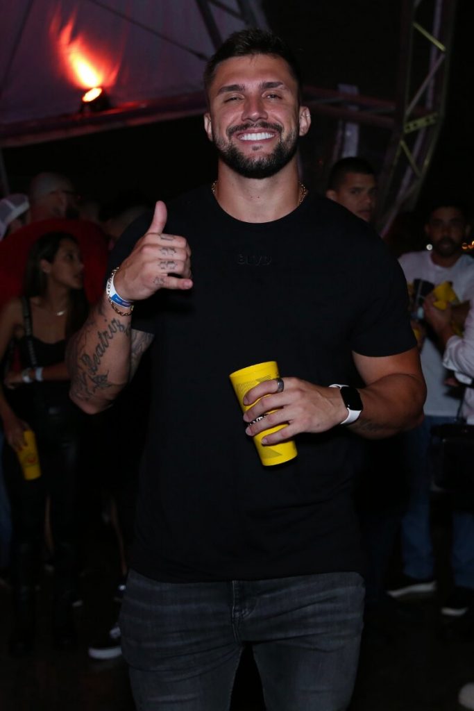 Arthur Picoli de boné preto e camisa preta,  fazendo sinal de positivo e segurando um copo amarelo