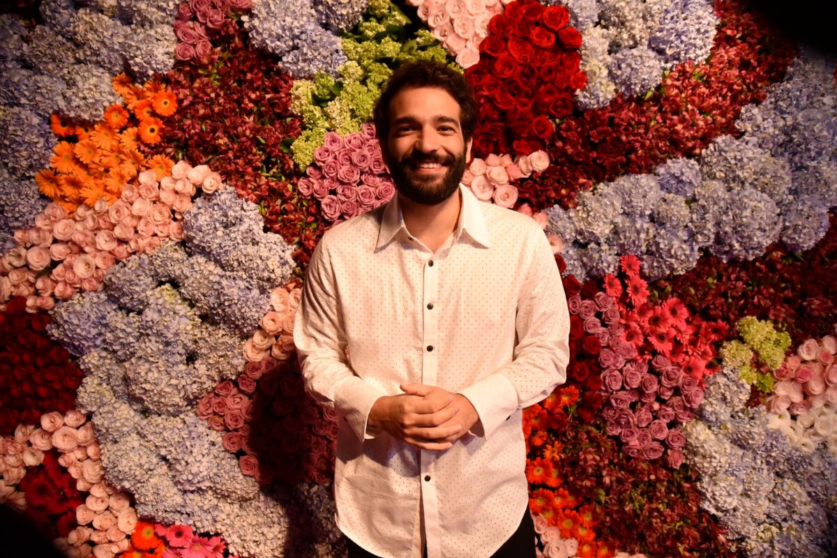 Humberto Carrão sorrindo, de camisa social branca, , diante de várias flores na parede