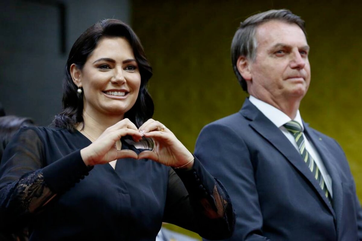 Michelle de preto, fazendo coração com as mãos e Jair Bolsonaro ao lado, de terno preto