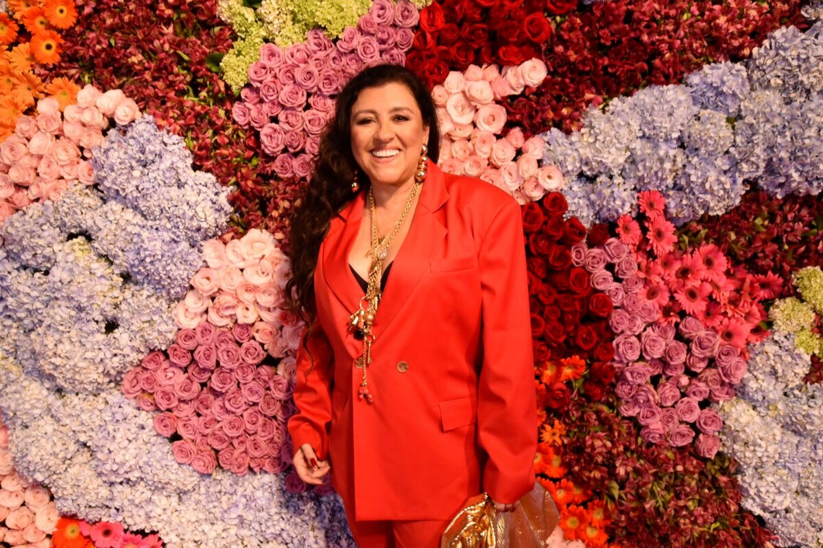 Regina Casé de calça e blazer vermelho segurando bolsa dourada, sorrindo, diante de várias flores na parede