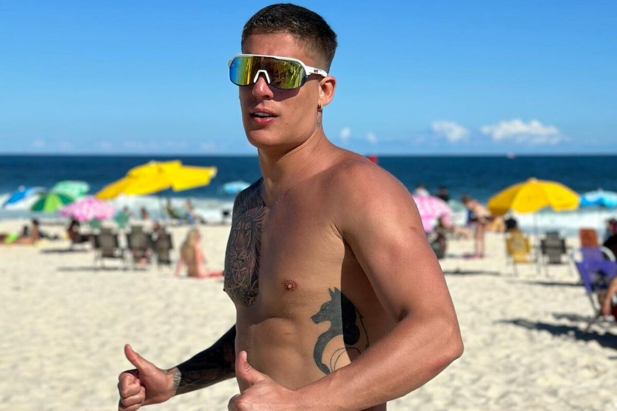 Tiago Ramos fazendo sinal de positivo de óculos escuros, sem camisa, em uma praia