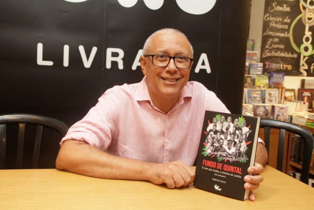 Marcos Salles de camisa social rosa,sentado, sorrindo, segurando livro