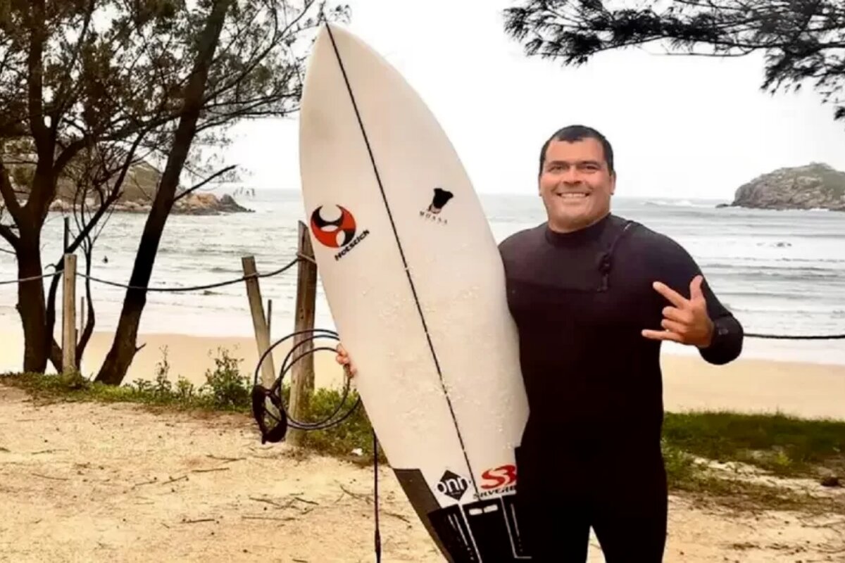 Marcio Freire de macacão preto de mergulho, segurando prancha de surf, fazendo sinal de hang-loose