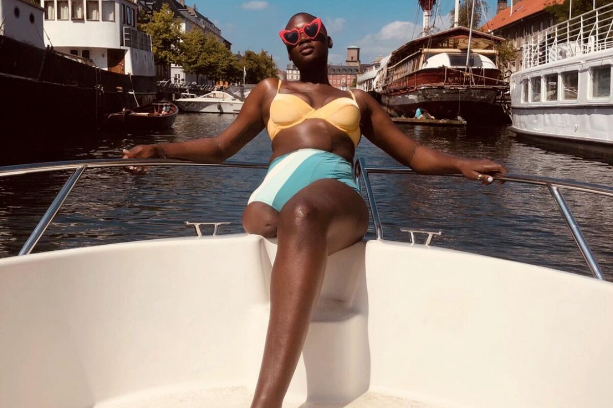 Modelo Mama Cax com uma perna só, de biquíni, sentada na proa de um barco, de óculos escuros