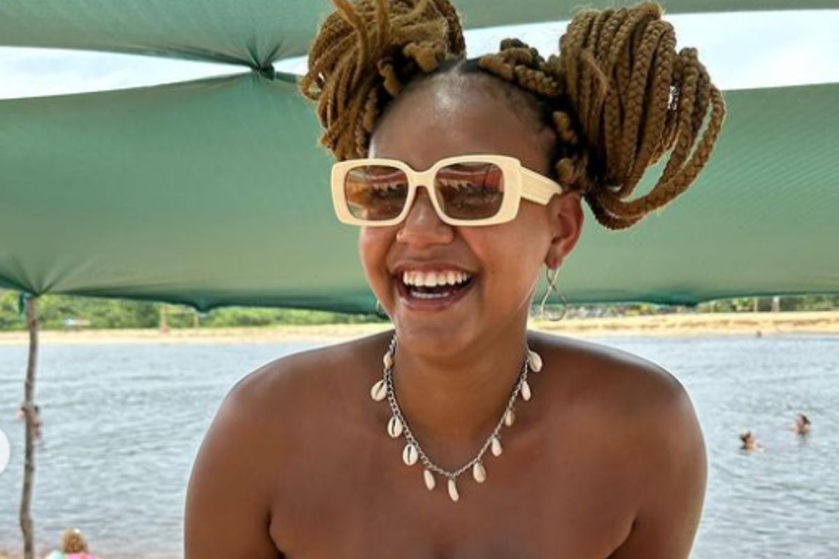Jessi Alves, com óculos de sol e cabelos presos, sorridente em foto, com praia ao fundo