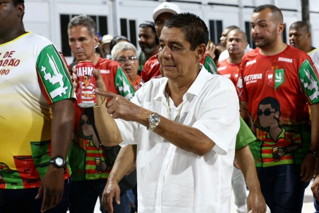 Zeca Pagodinho de camisa branca mostrando rótulo de cerveja, no ensaio técnico da Grande Rio na Sapucaí 