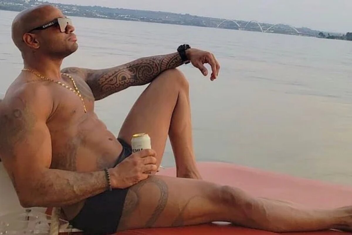 Cesar Black sentado de sunga, segurando lata de cerveja, com o mar ao fundo