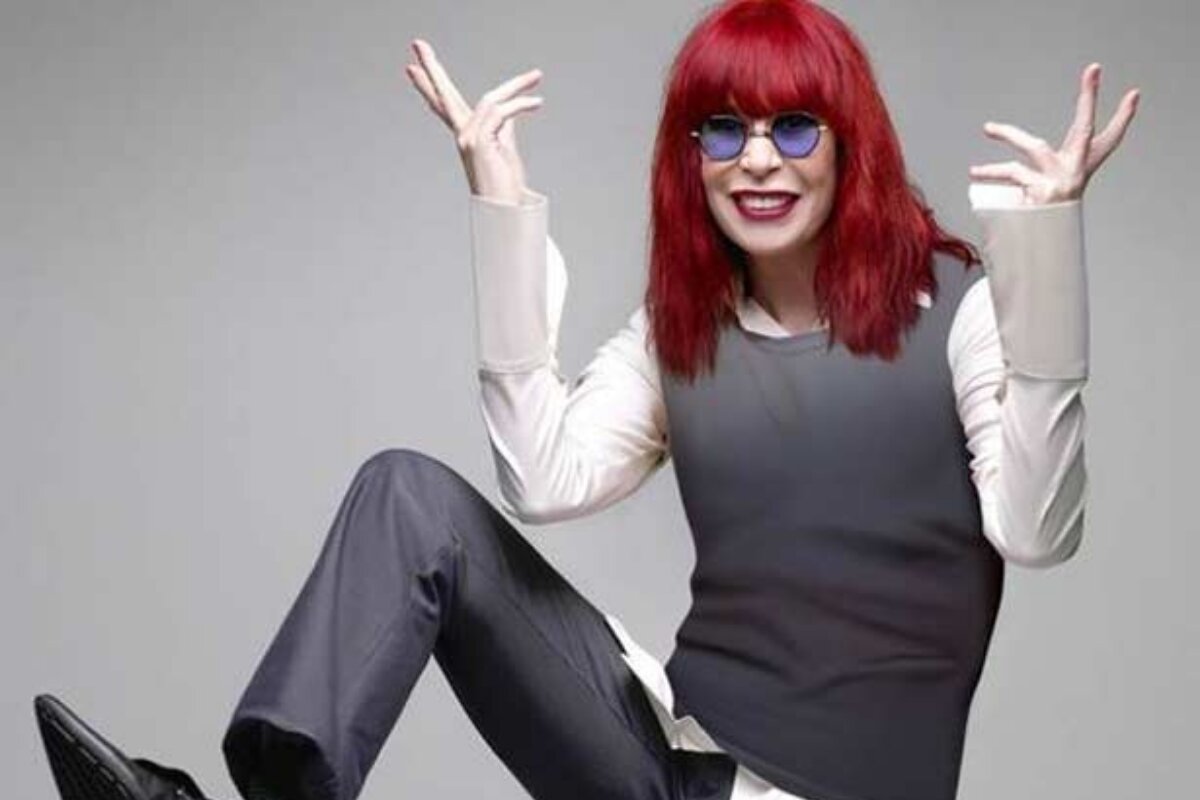 Rita Lee de cabelo vermelho, calça e colete cinza, camisa branca