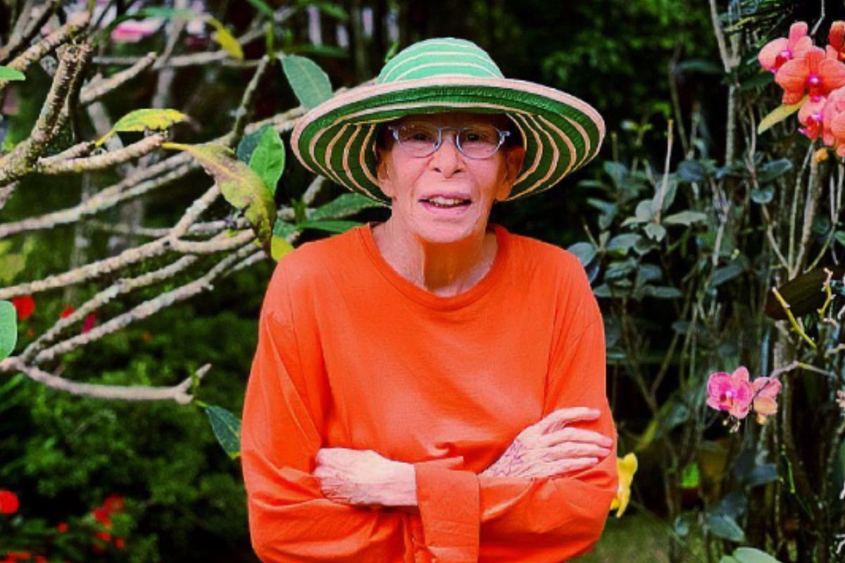 Rita Lee, sorridente , de chapéu e de braços cruzados no jardim