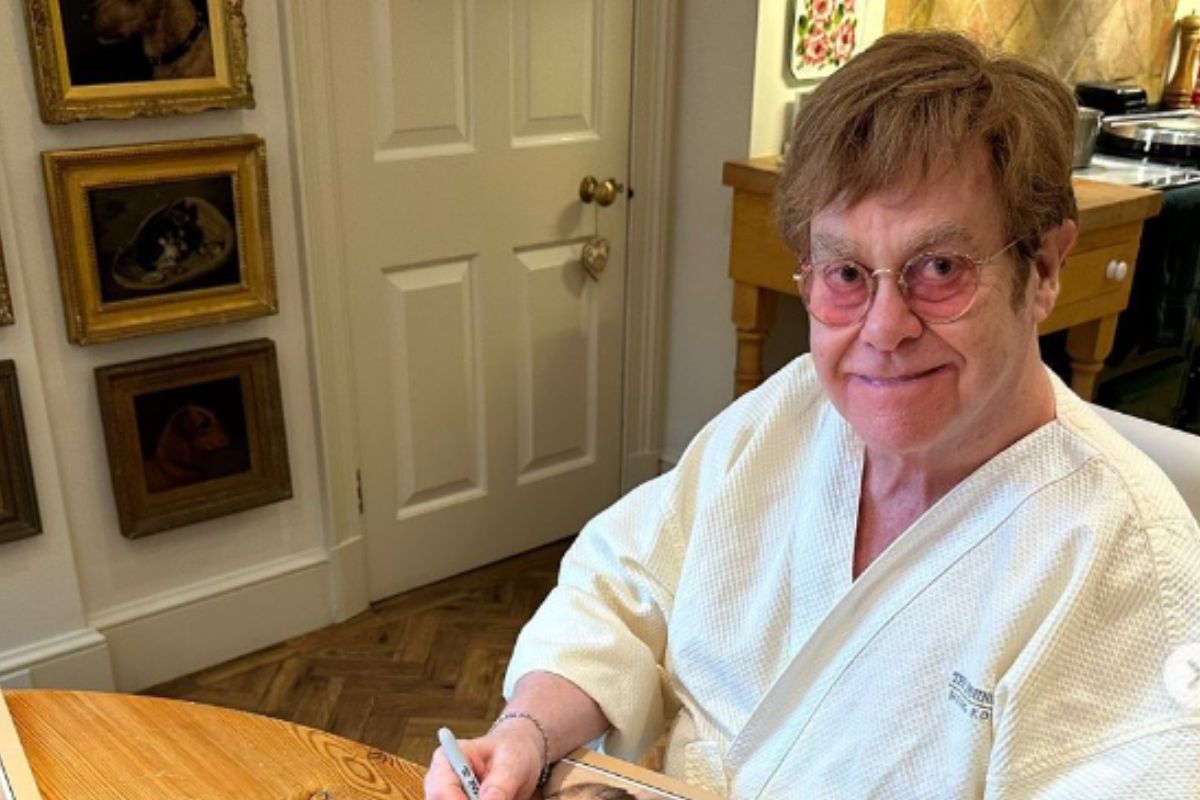 Elton John, de roupão, sentado em sua casa