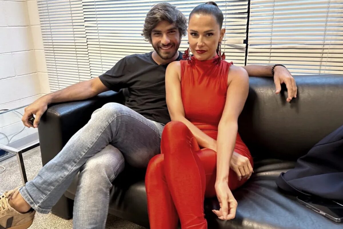 Hugo Moura e Deborah Secco de macacão vermelho sentados num sofá preto