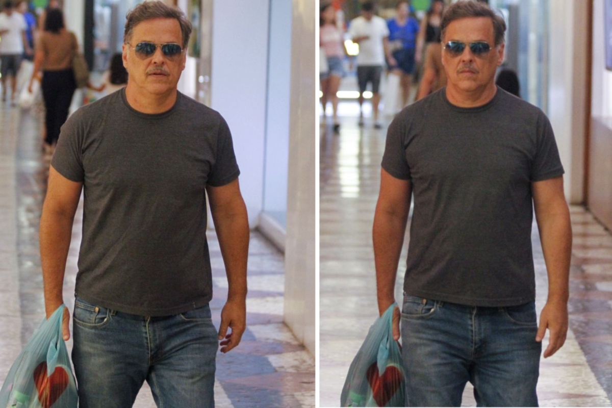 Guilherme Fontes de calça jeans e camisa cinza, óculos escuros, segurando sacolas, no corredor de um shopping