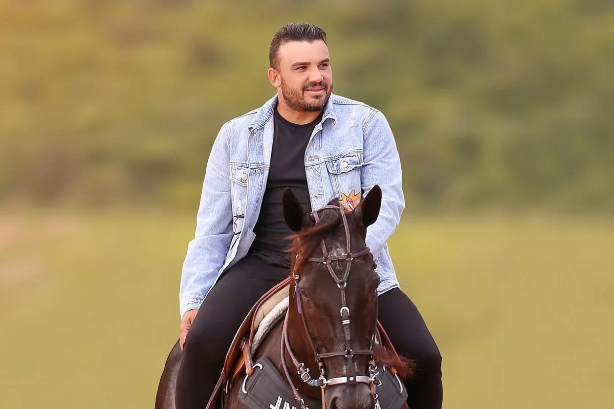Cantor de forró Júnior Vianna montado em um cavalo, de calça e camisa preta, blusa jeans