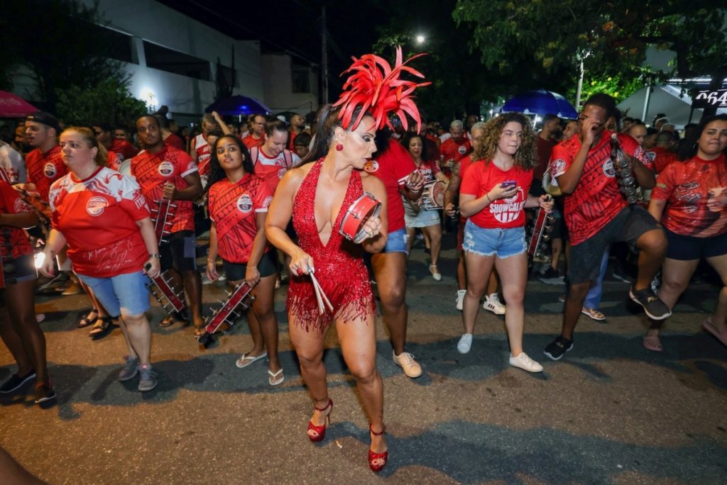 Viviane Araújo de adereço de penas na cabeça, body estilizado vermelho, tocando tamborim