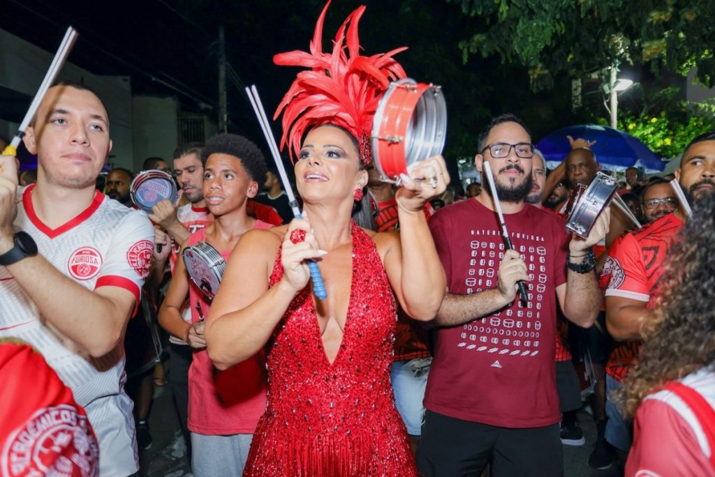 Viviane Araújo de adereço de penas na cabeça, body estilizado vermelho, tocando tamborim